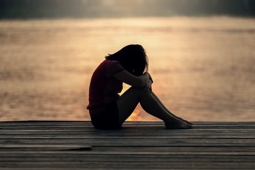 Girl sits alone with her head down in silhouette near a body of water.