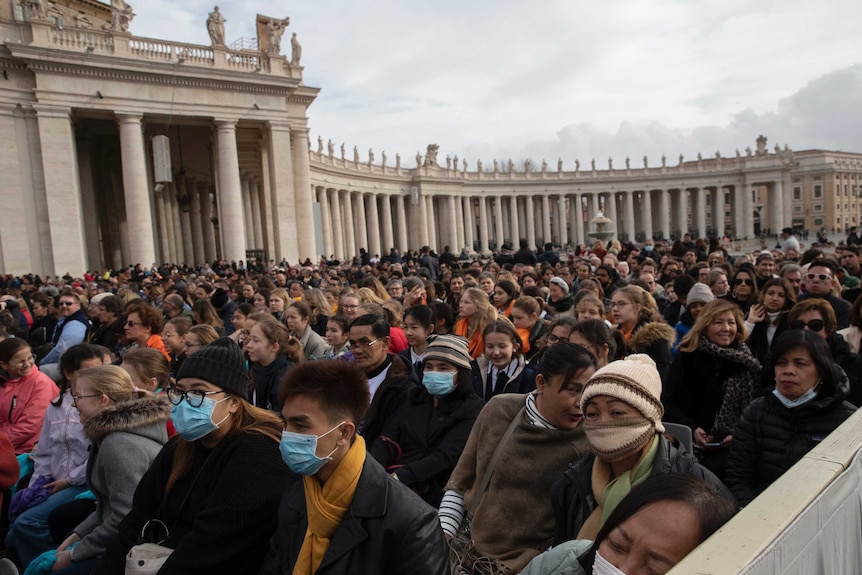 A crowd of people in winter clothes, with some wearing face masks, stand together on a cloudy day.