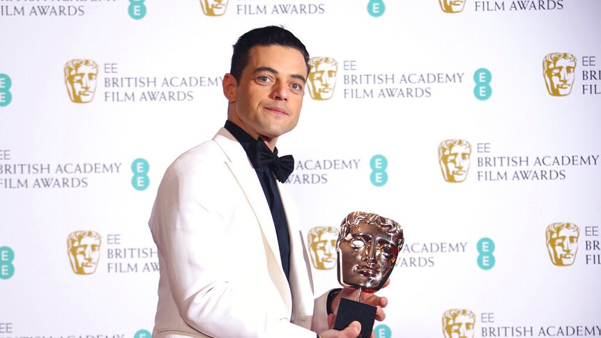 Remi Malek holds a BAFTA statue while wearing a white suit jacket with a black bow tie.