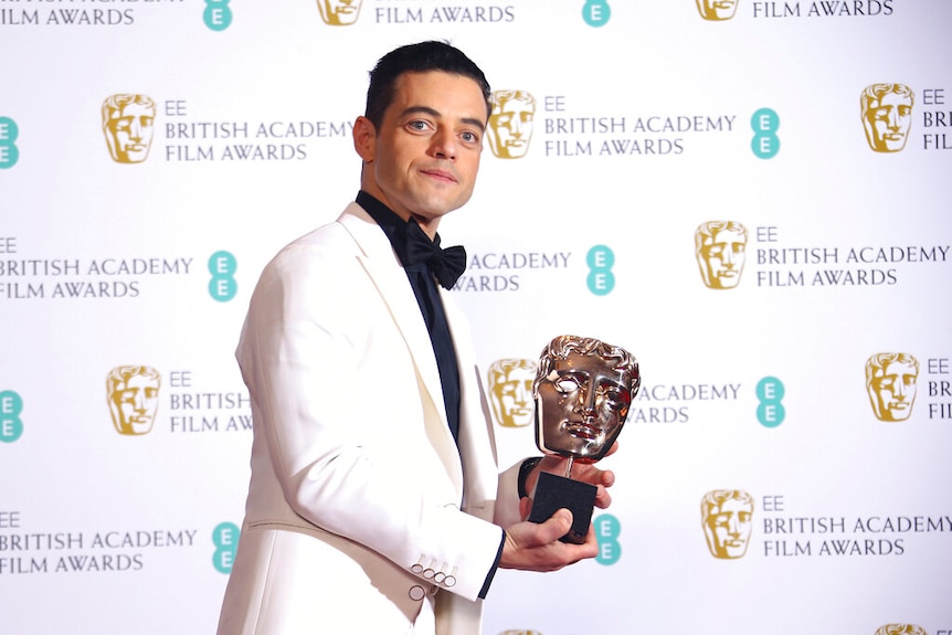 Remi Malek holds a BAFTA statue while wearing a white suit jacket with a black bow tie.