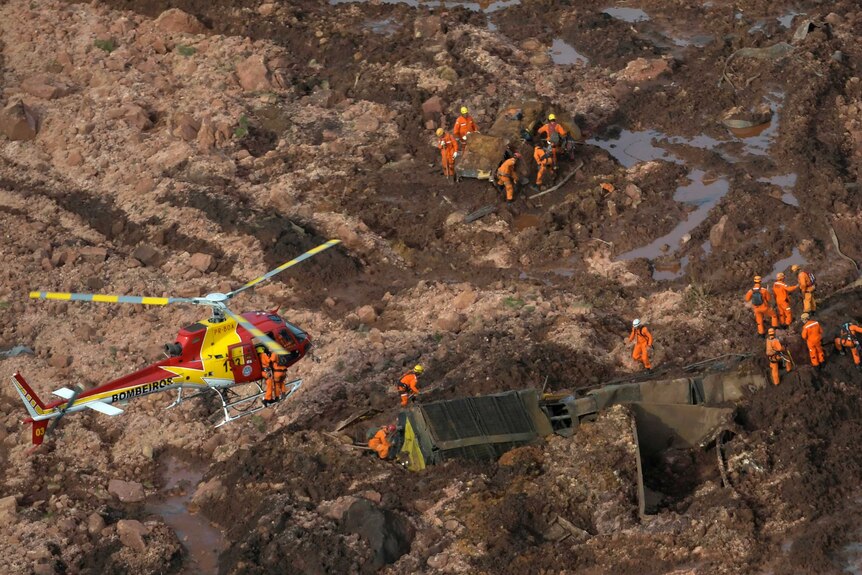 An aerial photo shows a field of brown sludge with tiny orange-clad rescue workers as a yellow and red helicopter flies ahead.
