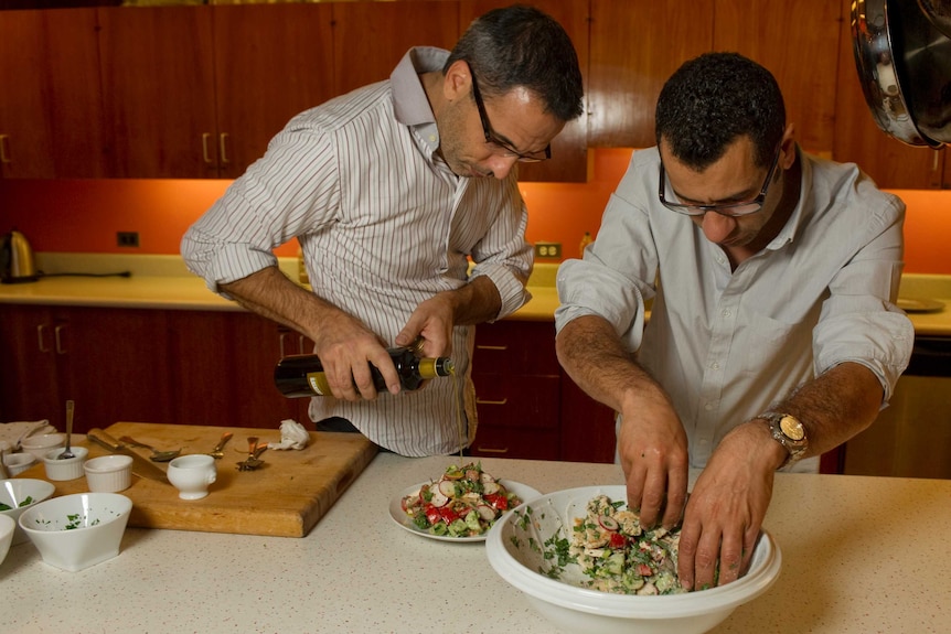 Two men prepare a salad; one drizzles olive oil over vegetables as the other mixes a bowl with his hands.