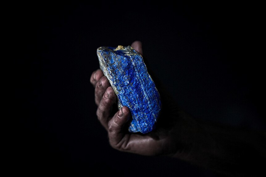 A darkened hand holding a piece of Lapis lazuli rock in front of a black background.