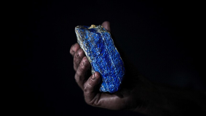 A darkened hand holding a piece of Lapis lazuli rock in front of a black background.
