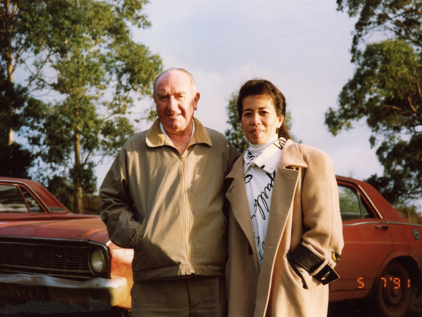 An older Anglo man and a young Filipina wife pose for a photo in front of a red vintage car.