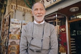 A man who appears to be in his late 70s stands outside his business in the bustling bazaar