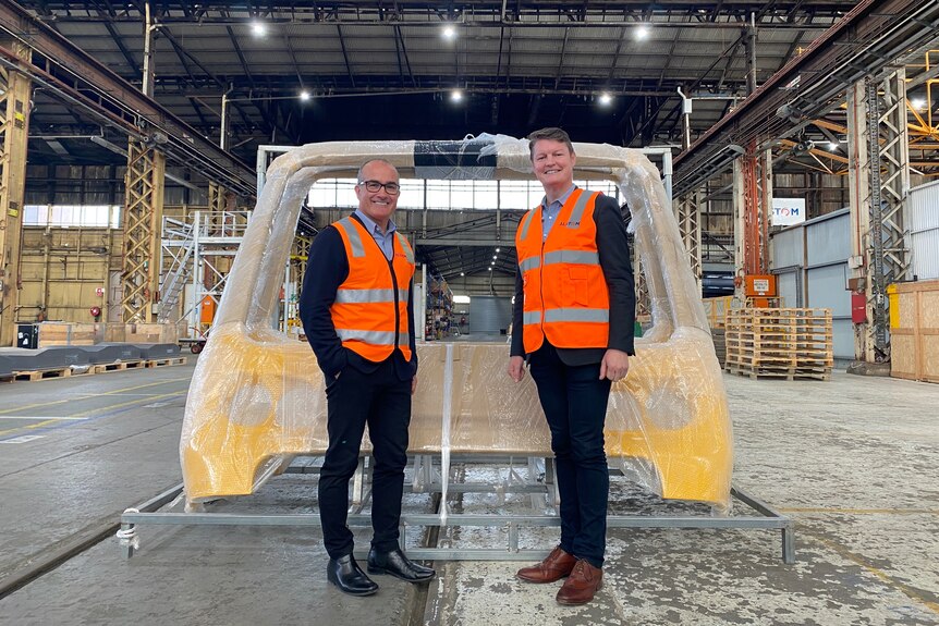 Two men in orange high vis stand in front of parts for a train in factory.