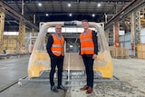 Two men in orange high-vis stand in front of parts for a train in factory.