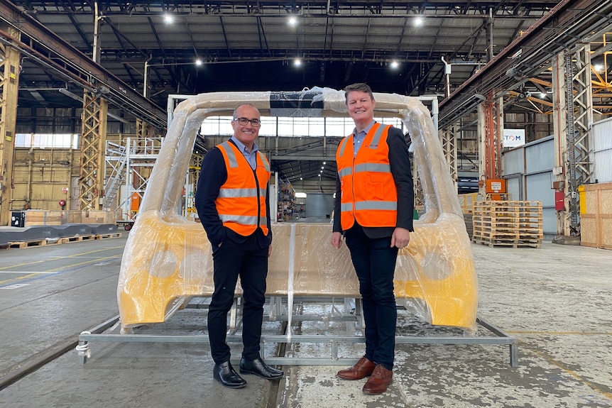 Two men in orange high vis stand in front of parts for a train in factory.