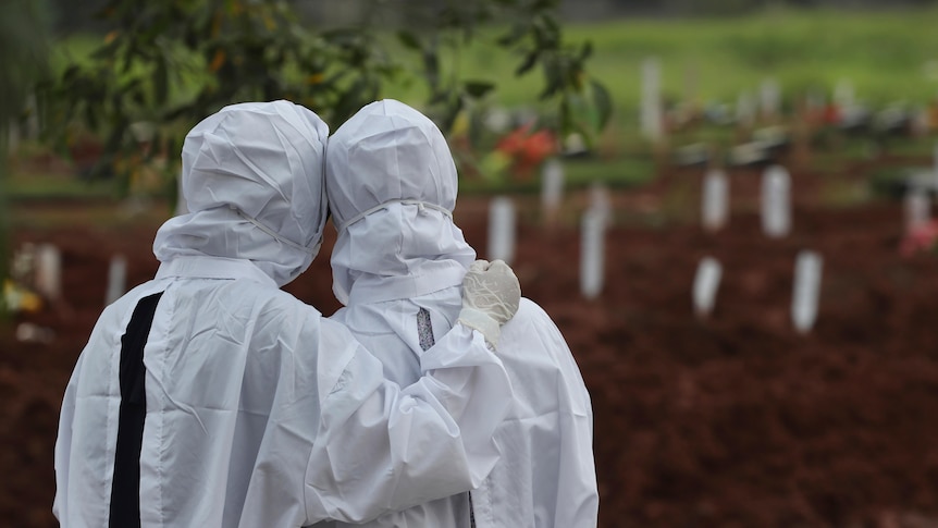 Two people wearing white protective suits console each other in a cemetery