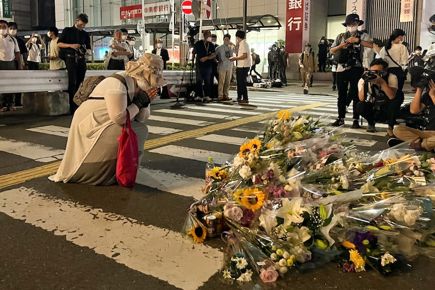 A woman kneels on the street with her head in her hands beside some flowers and photographers.