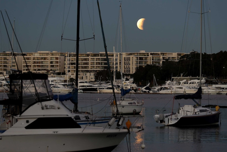 The moon is seen over apartments and boats on the harbour