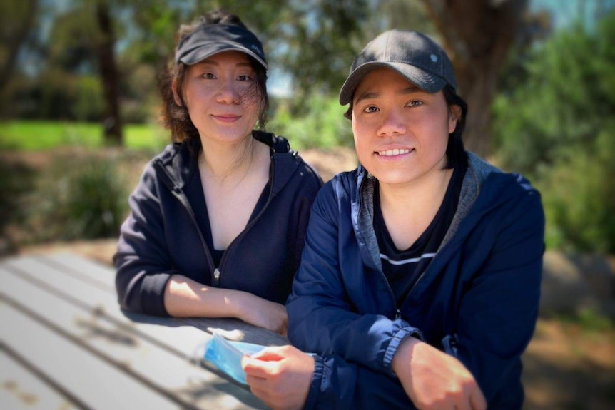 Two women sit in a park at a bench smiling, they wear hats and active wear clothing.