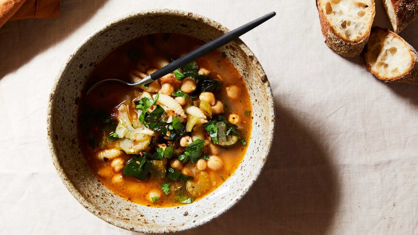 A bowl of pasta, chickpea and spinach soup with a napkin and crusty bread nearby, a hearty family meal.