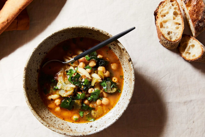 A bowl of pasta, chickpea and spinach soup with a napkin and crusty bread nearby, a heart family meal.