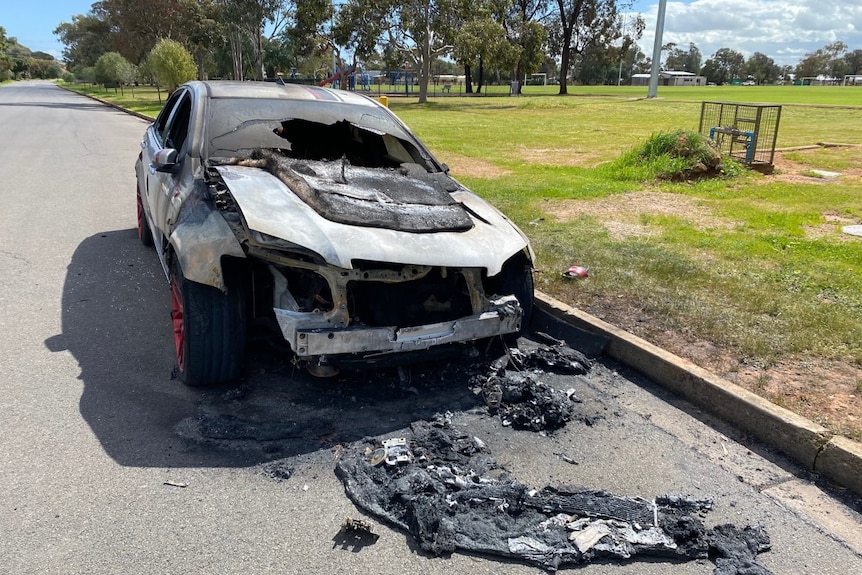 A burnt silver car surrounded by melted plastic and broken glass, parked next to a grassy park