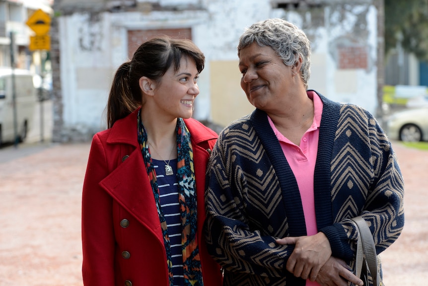 A 20-something woman and an older Black woman walk arm in arm on a Sydney street, smiling at each other