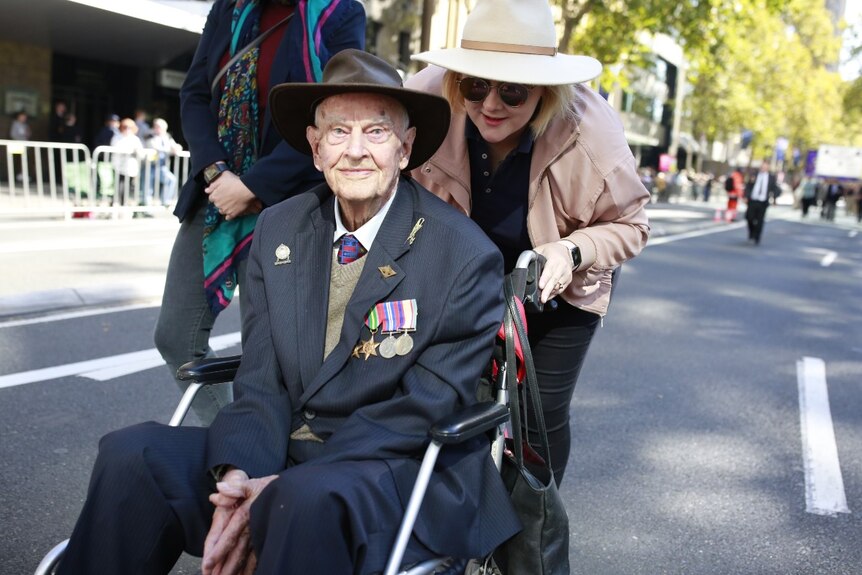 An elderly man wearing medals is pushed in a wheelchair by a younger woman.