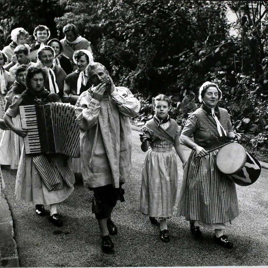 a black and white image of a troupe of folk musicians walking towards the camera, carrying handheld instruments