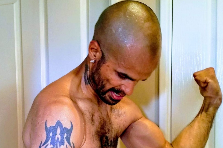 A shirtless man with a big tattoo on his right arm flexing his muscles for the camera