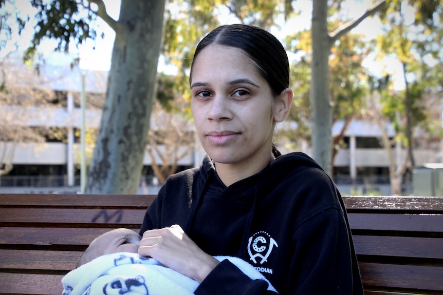 A woman sits on a park bench, looking at a camera, holding her baby.