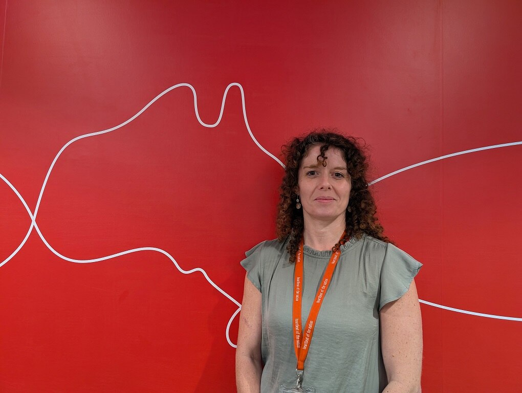 A woman with brown curly hair stands in front of a red backdrop.