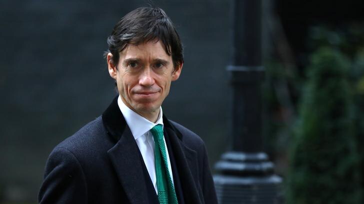 Rory Stewart smiles slightly as he looks to the right. He wears a dark overcoat and a suit with a green tie.