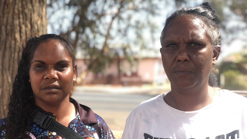 Two Aboriginal women face the camera with a suburban street and a tree in the background.
