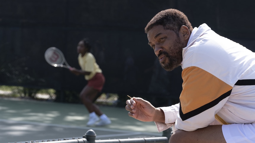 A middle-aged African American man sits courtside grimly watching a teenage girl play tennis, holding a toothpick
