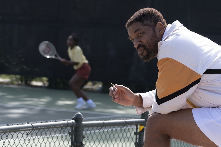 A middle-aged African American man sits courtside grimly watching a teenage girl play tennis, holding a toothpick