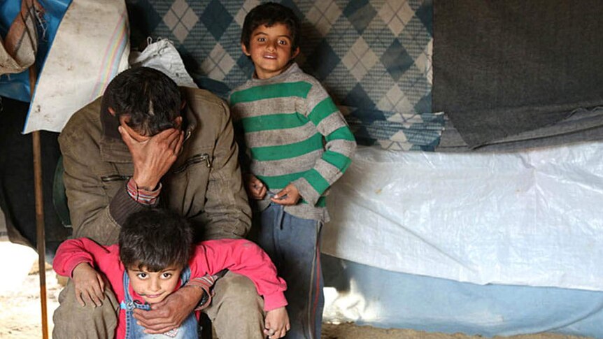A Syrian man holds his head in his hands as he sits with two small children next to him.