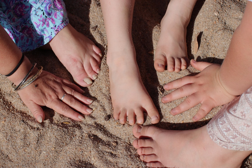 Close up of mum's hands and feet with her children's smaller hands and feet in sand