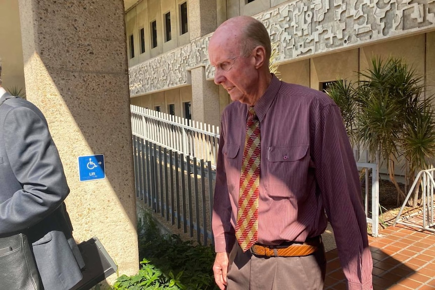 A frail elderly man wearing a button down shirt and tie leaving a courthouse.