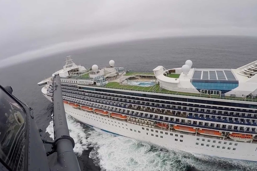 A cruise ship sitting in water with a helicopter hovering above.