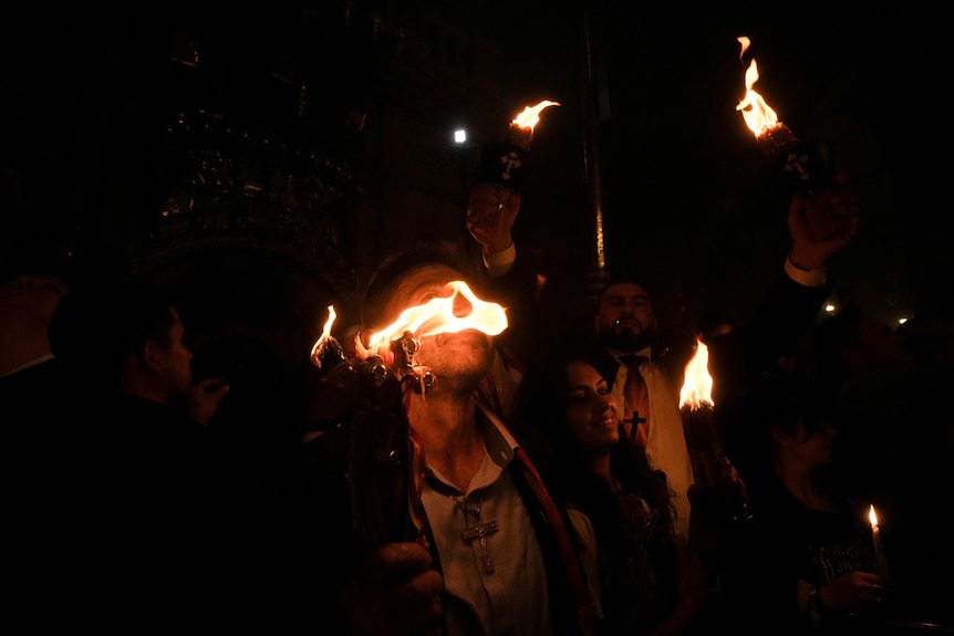 A bright orange flame burns near a person's face as people stand together holding candles.