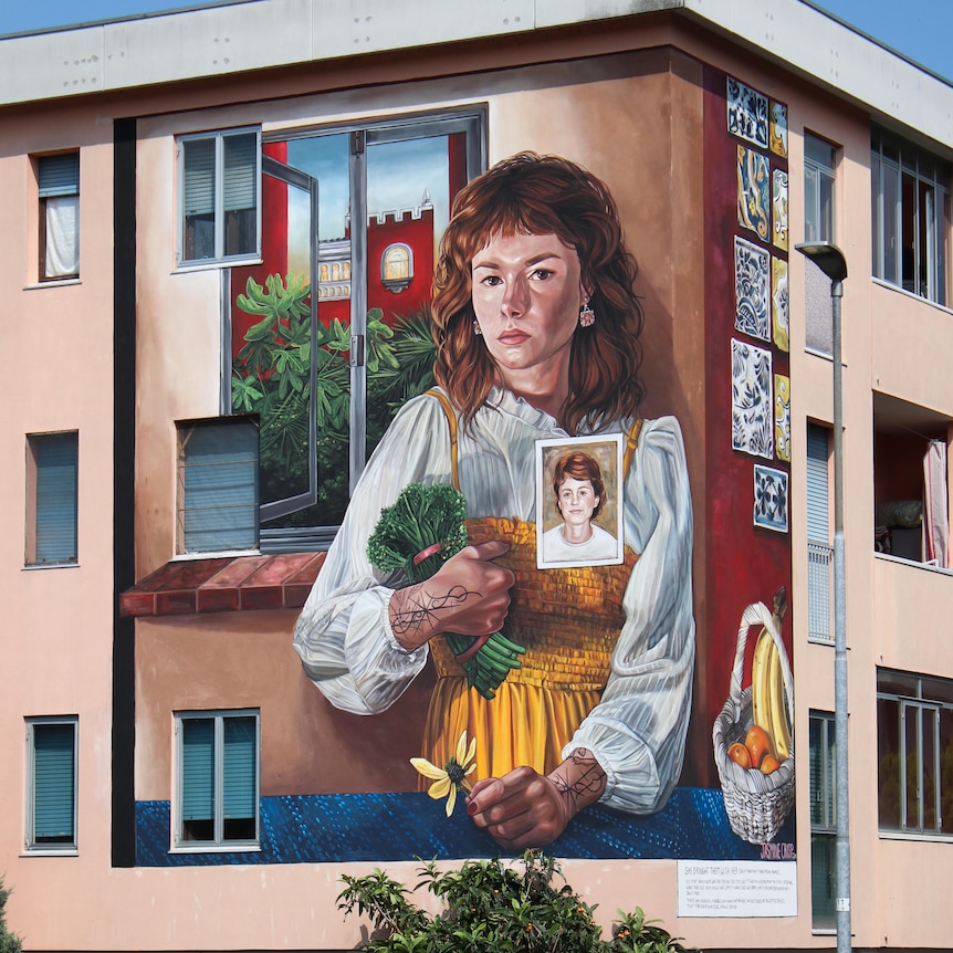 large mural of woman with curly red hair holding broccoli on the side of a three story building