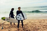 Man and woman in wetsuits hold surf boards and look out at waves