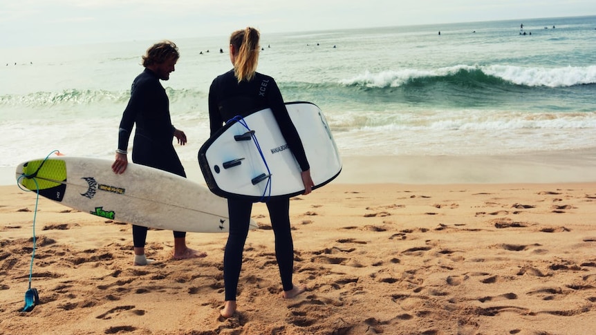 Man and woman in wetsuits hold surf boards and look out at waves