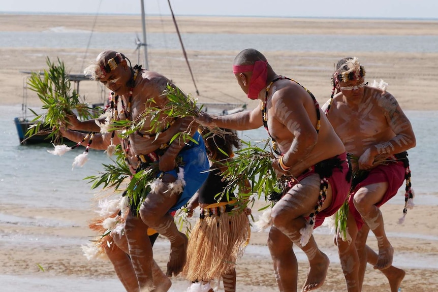 A group od Aboriginal men dance on a beach. They are wearing traditional dress and are painted with white paint.