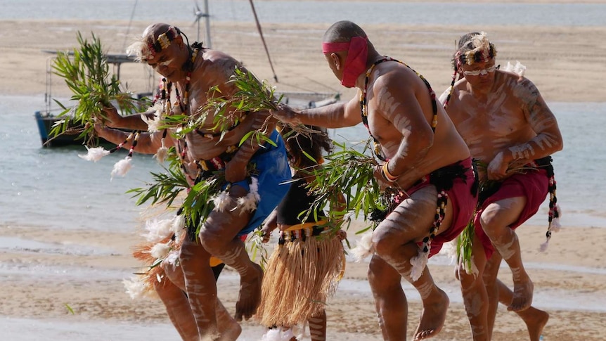 A group od Aboriginal men dance on a beach, wearing traditional dress and are painted with white paint.
