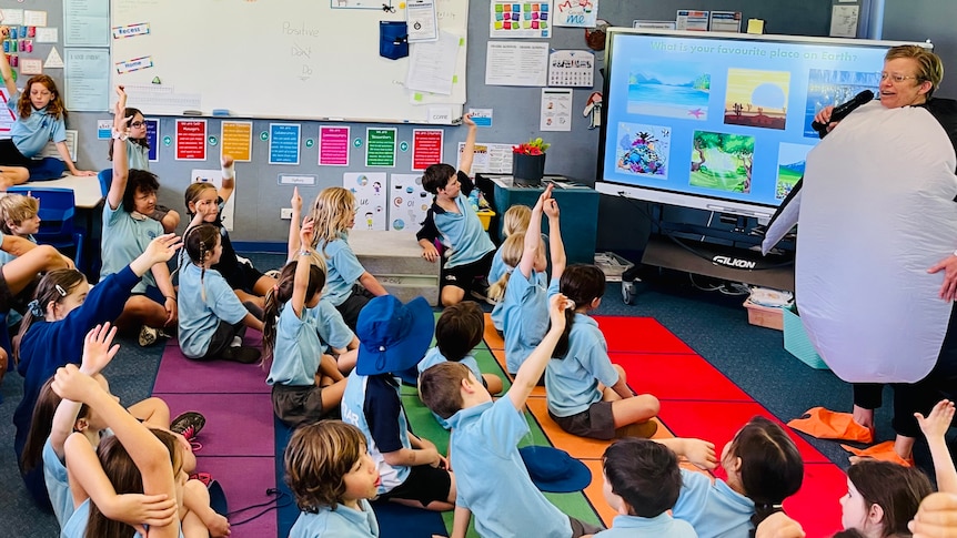 A woman with cropped blonde hair and glasses dressed as a penguin, speaking to a class of children