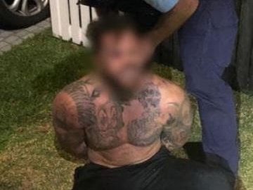 A shirtless, heavily tattooed man sits on grass while he is arrested by a NSW police officer.