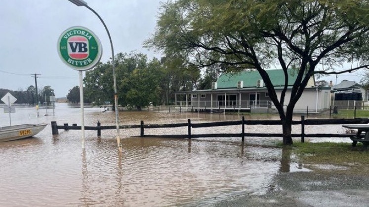 Brown flood waters encroaching on a wooden fence and home. tinny on the side and large round green sign with 'vb' on a tall poile