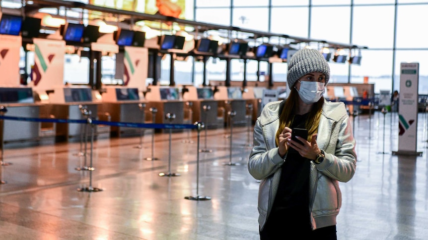 A woman stands in an empty airport terminal wearing a face mask