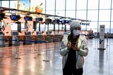 A woman stands in an empty airport terminal wearing a face mask
