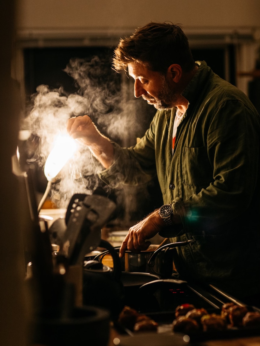 Tim Thatcher sprinkles something over a pan on the stove, as light illuminates steam rising from the pans.