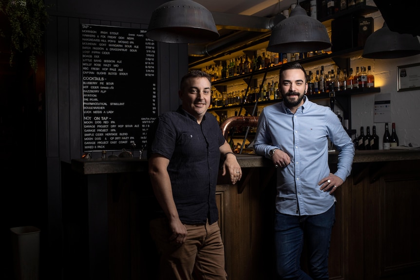 Two men stand at a bar and look at the camera