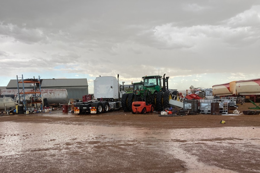 Machinery, trucks, and shelves in the open air, on muddy, wet ground with a dark sky above.