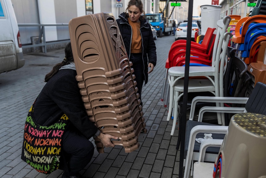 A man is bent down lifting up a stack of chairs and a woman is watching on. 