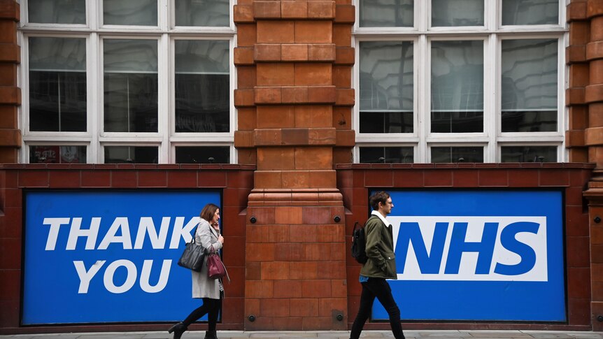 Two people walk in front of a large banner that reads "Thank you NHS"